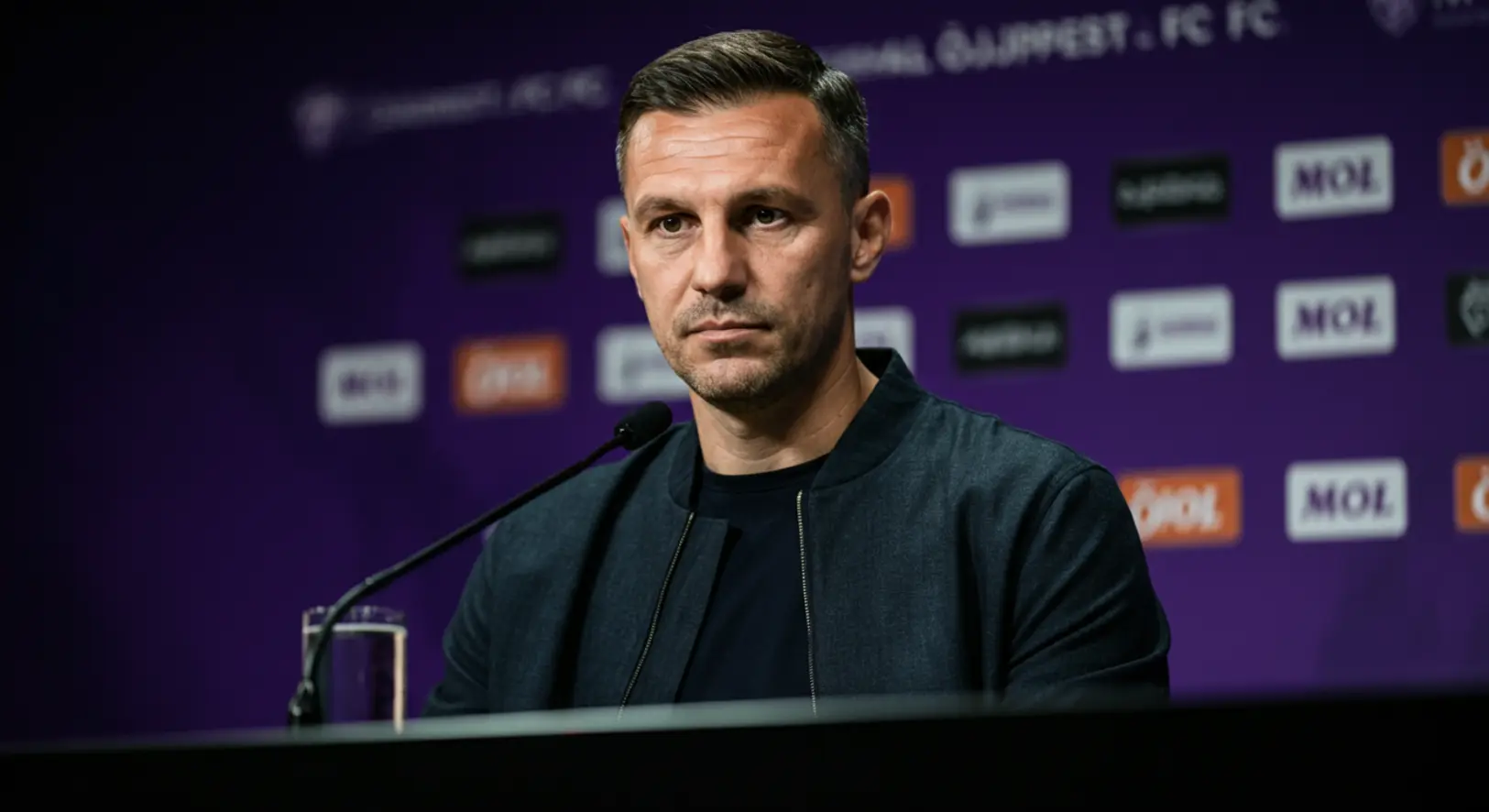 Dárdai Pál komoly tekintettel beszél az Újpest FC sajtótájékoztatóján, háttérben a klub lila-fehér színeivel és a MOL logójával.