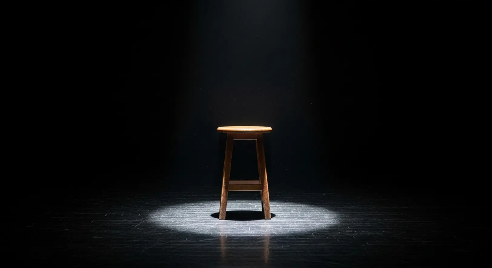 Egyetlen üres fa hokedli áll egy sötét színházi színpad közepén, felülről érkező éles reflektorfényben megvilágítva, szimbolizálva a hiányt.