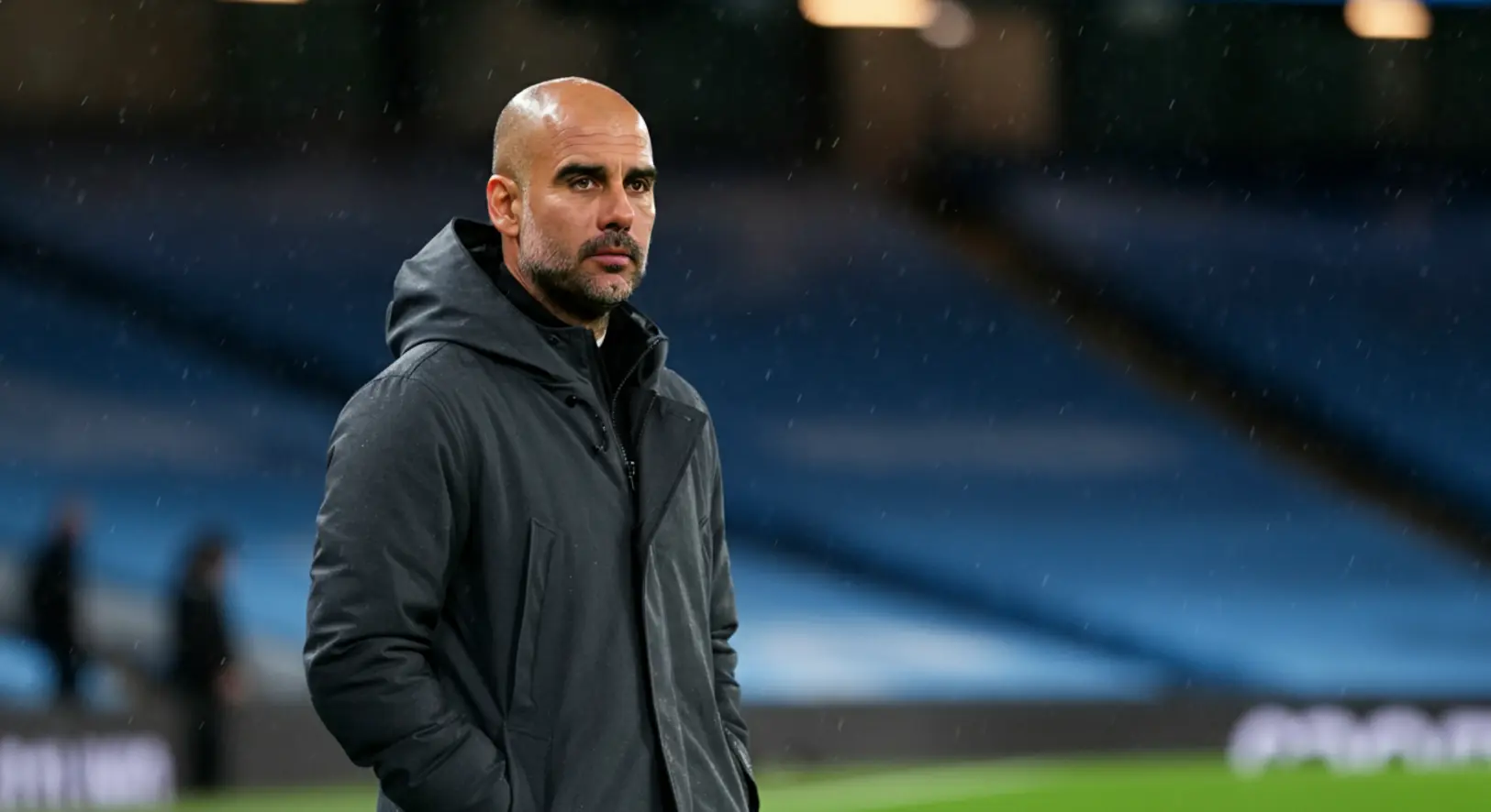 Pep Guardiola, a Manchester City menedzsere sötét kabátban, esőben, intenzív arckifejezéssel figyeli a játékot az Etihad Stadion partvonaláról egy esti mérkőzésen.